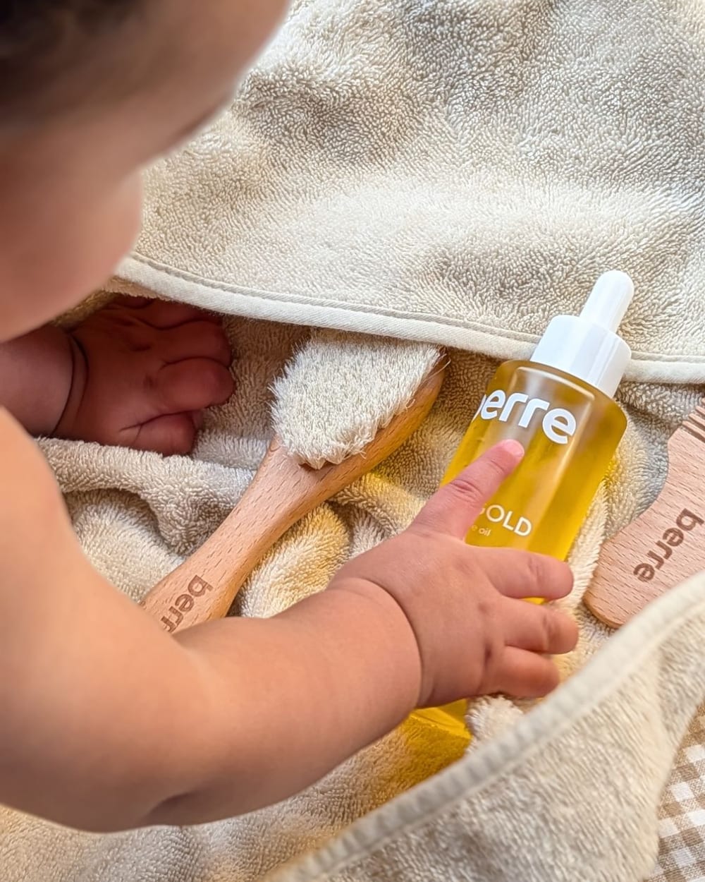 Baby reaching for Berre Liquid Gold natural baby oil beside the Berre baby brush and comb on an organic cotton hooded towel
